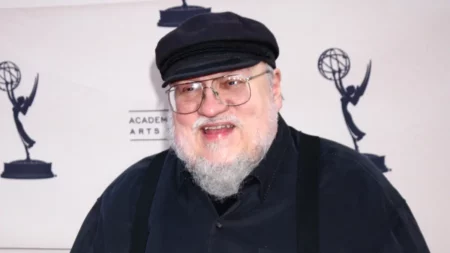 Criador de Game of Thrones desabafa sobre depressão após meses afastado: ‘Avassalador’ George R. R. Martin sorri durante evento, usando boné preto, óculos e barba branca, após reaparecer publicamente em meio a relato sobre depressão e envelhecimento.