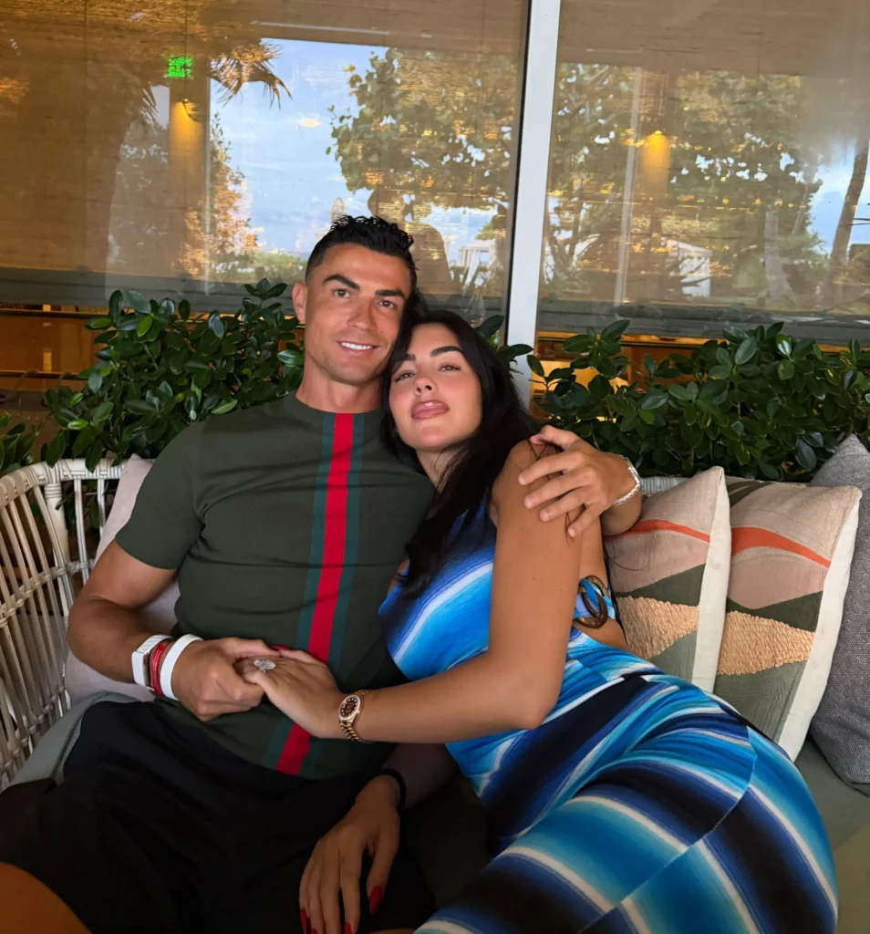 Cristiano Ronaldo e Georgina Rodríguez sentados juntos em um sofá, de mãos dadas e relaxados.