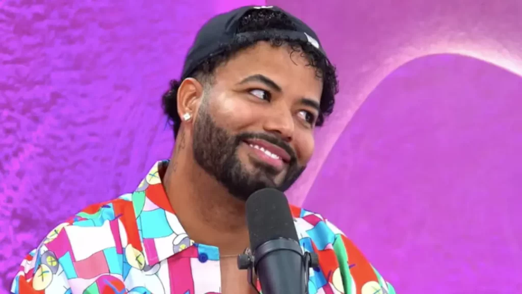 Hytalo Santos aparece sorrindo diante de um microfone, usando boné e camisa estampada, em frente a um fundo roxo.