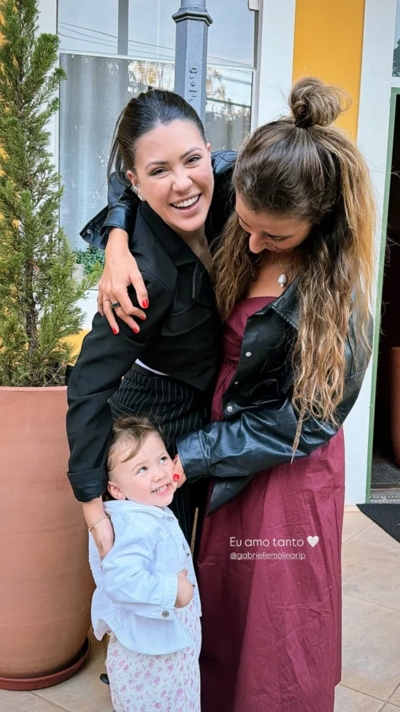 Nina Forlin, com amiga e uma criança aparecem sorrindo em frente a uma hospedagem durante o passeio.