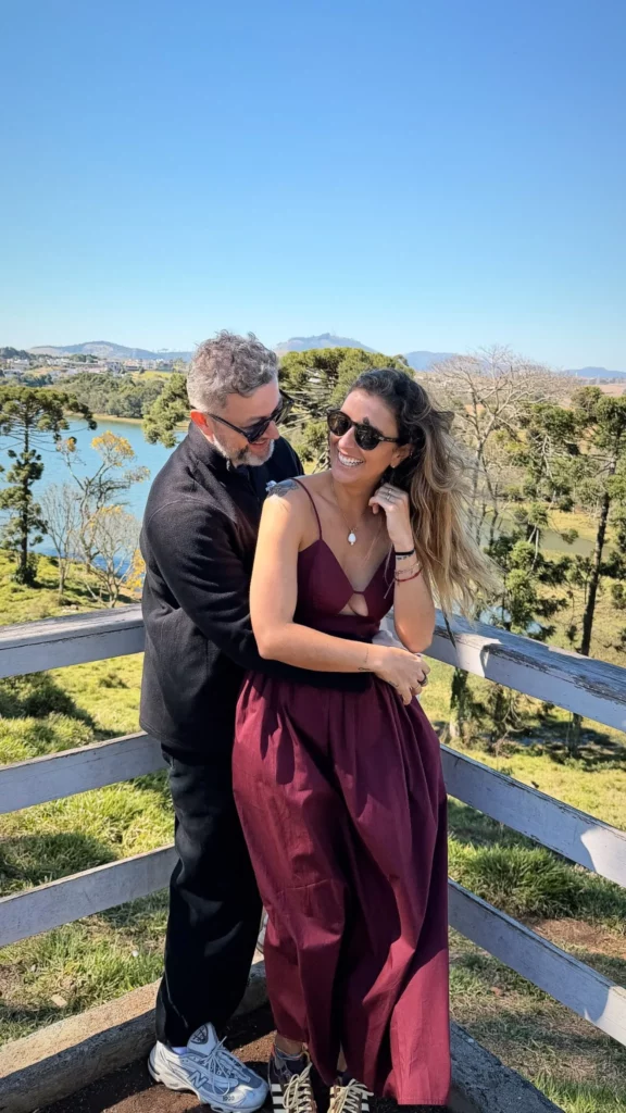 Lucas Lima abraça Nina Forlin em um mirante com vista para um lago durante viagem romântica.