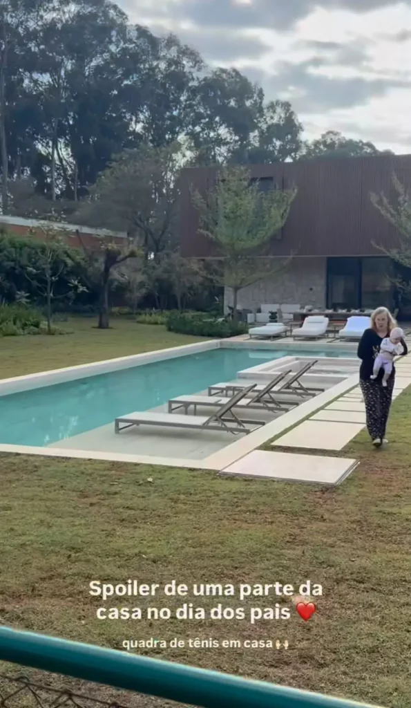 Mulher aparece com uma criança ao lado da piscina da nova mansão, com espreguiçadeiras e área verde ao redor.