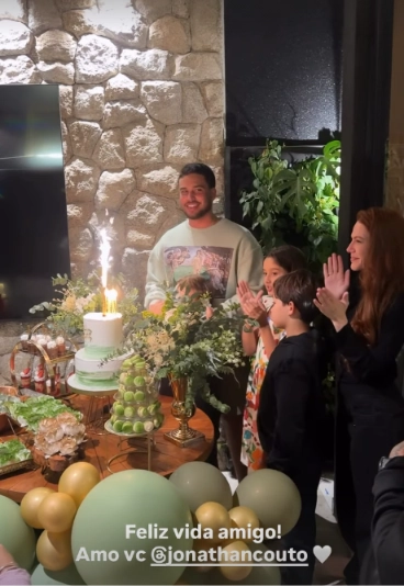 Jonathan Couto sorri diante do bolo de aniversário enquanto Sarah Poncio, ao lado dos filhos, aplaude durante o parabéns