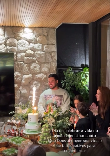 Jonathan Couto sorri diante do bolo de aniversário enquanto Sarah Poncio, ao lado dos filhos, aplaude durante o parabéns