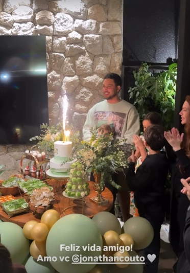 Jonathan Couto sorri diante do bolo de aniversário enquanto Sarah Poncio, ao lado dos filhos, aplaude durante o parabéns