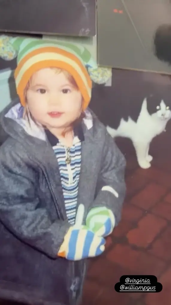 Virginia criança sorrindo levemente, usando touca colorida com pompons, casaco de frio e luvas listradas.