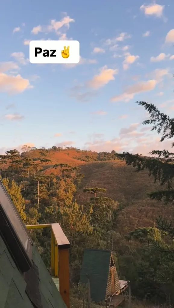 Vista da varanda de um chalé para colinas florestadas ao pôr do sol, com um balão de texto 'Paz' e um emoji de paz.