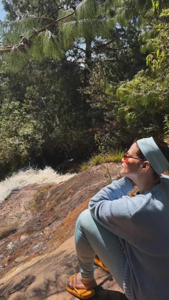 Thais Carla sentada em uma pedra ao sol, de perfil, olhando para uma cachoeira ao fundo.