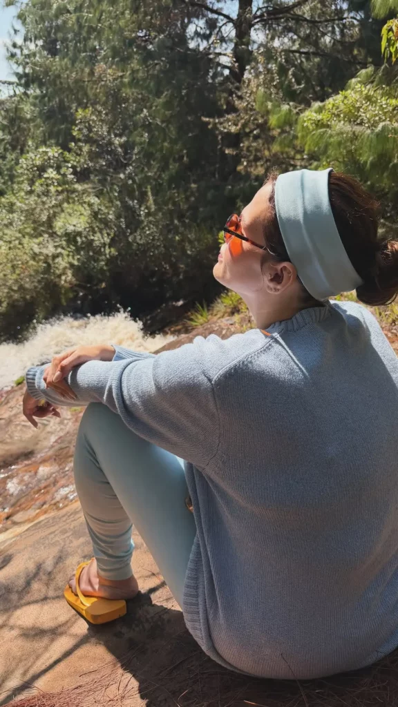 Thais Carla sentada em uma pedra ao sol, de perfil, olhando para uma cachoeira ao fundo.