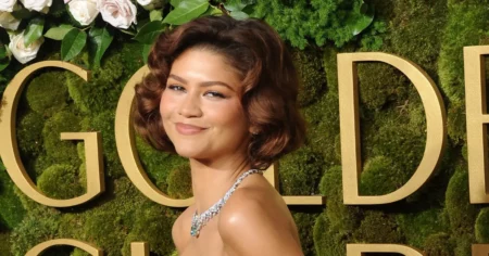 Zendaya posa em evento com cabelo curto e ondulado, colar de pedras e vestido sem alças, diante de um painel verde decorado com flores e letras douradas