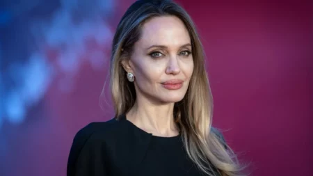 Angelina Jolie posa para foto com cabelos longos e vestido preto diante de fundo colorido.