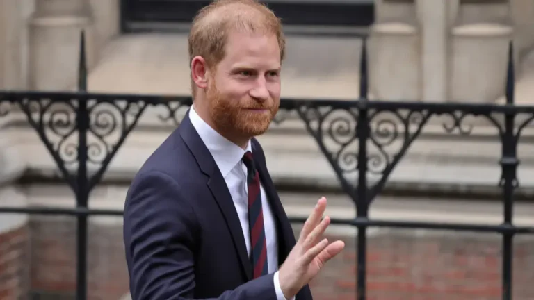 Príncipe Harry aparece de terno e gravata, acena enquanto caminha diante de um prédio.