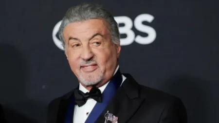 Sylvester Stallone aparece de terno preto e gravata borboleta em evento, sorrindo diante de um painel com logotipo.