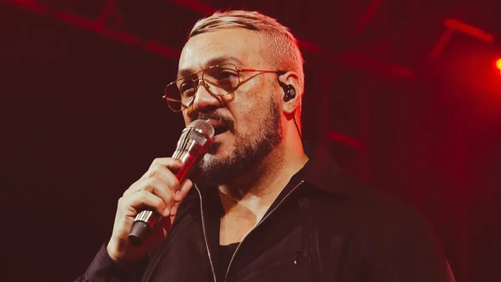 Belo enfrenta nova cobrança judicial e pode ter R$ 284 mil retidos; entenda Belo aparece de óculos e roupa preta durante apresentação musical, segurando um microfone sob iluminação vermelha.