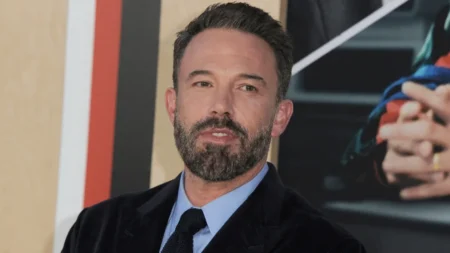 Ben Affleck aparece de terno preto e camisa azul em evento, olhando para a câmera.