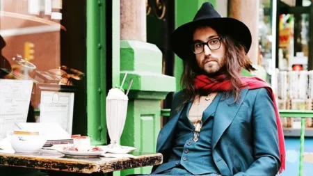 Filho de John Lennon revela divisão da herança do pai: ‘Diferença era insignificante’ Sean Lennon usa chapéu preto, óculos e cachecol vermelho enquanto está sentado à mesa de um café.