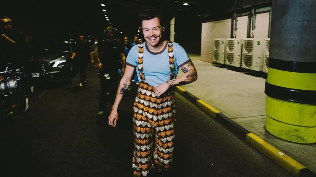 Harry Styles discute com caçador de autógrafos em Nova York; veja Harry Styles sorri em preparação pré-show, usando camiseta azul e jardineira estampada.