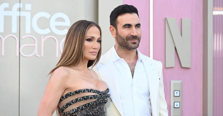 Jennifer Lopez posa ao lado de Brett Goldstein em evento, usando vestido preto com detalhes brilhantes, enquanto o ator veste terno branco
