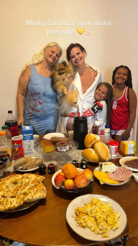 Solteira, Thais Carla dança de roupão ao ganhar surpresa no aniversário de 35 anos 1 Thais Carla, de roupão branco, posa ao lado da mãe, das filhas Maria e Eva e do cachorro durante o café da manhã de aniversário. A mesa está repleta de pães, bolos, frutas e outras comidas. Acima deles, a frase “Minha família é meu maior presente”.