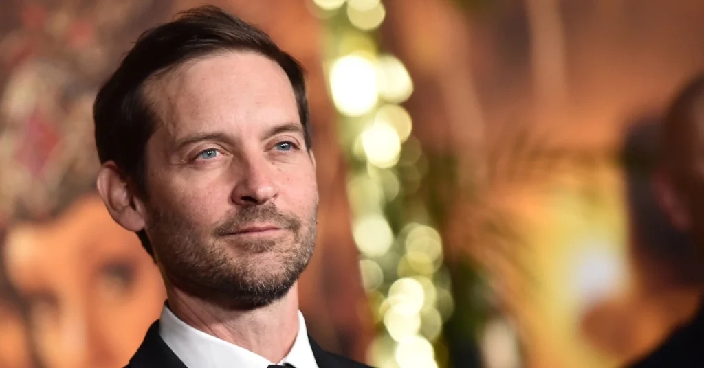 Tobey Maguire casa a ex-mulher com herdeiro bilionário; entenda Tobey Maguire aparece de terno preto e gravata, com barba curta, durante evento com iluminação desfocada ao fundo