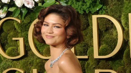 Zendaya posa com vestido elegante e colar de diamantes diante de um painel verde com letras douradas.